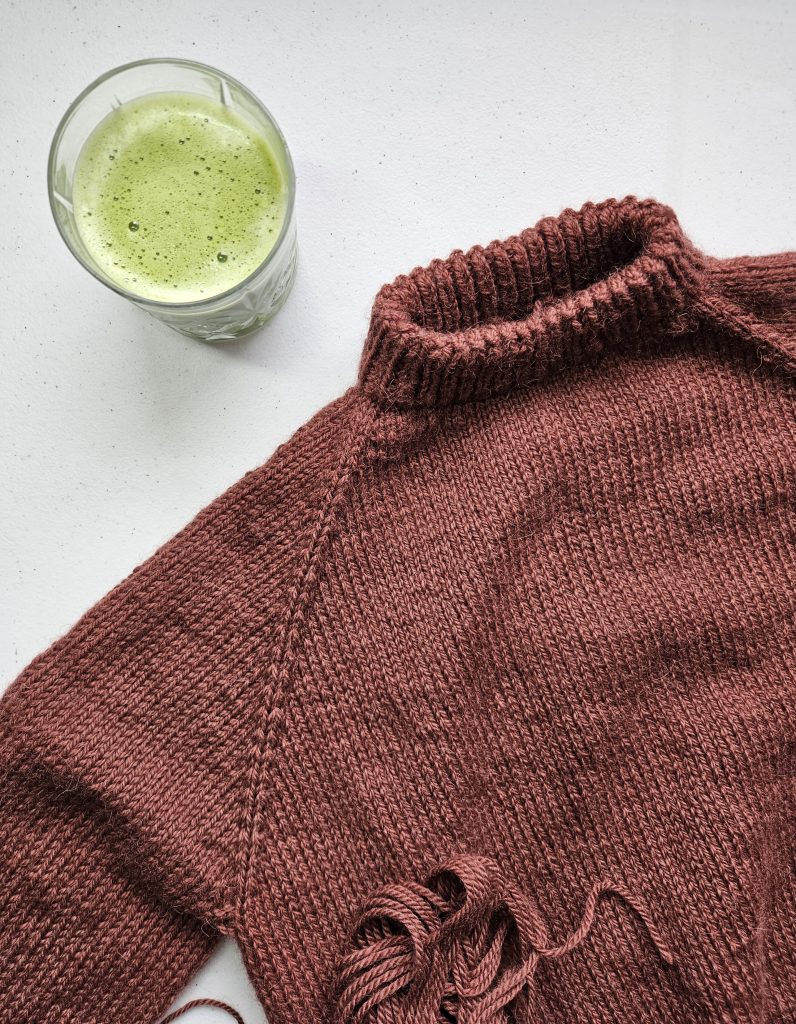 A burgundy handknit sweater laid on a table next to a glass of matcha latte.
