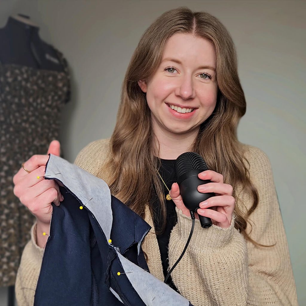 A photo of the blog author, Amy J Stitches, holding up a work in progress and a microphone. In the background you see a dressform wearing a green, floral dress