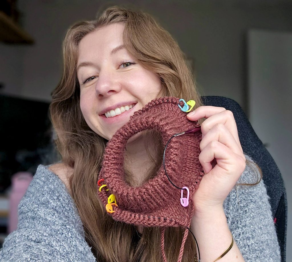 Amy J Stitches holding up the finished collar of her first knitting project, the step by step sweater. The sweater is a deep burgundy colour, and Amy is smiling as she holds it.